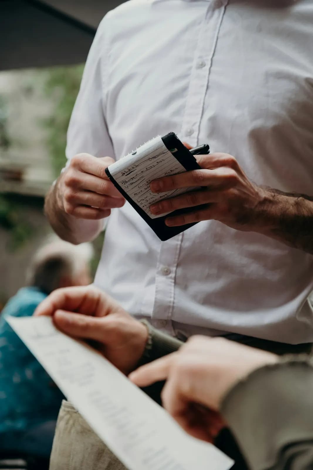 نادل بقميص أبيض يدوّن طلباً في دفتر صغير، وأمامه عميل يمسك قائمة الطعام